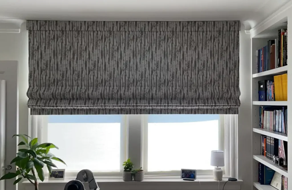 Gray Roman shades by Window Treatments North Carolina partially cover two windows in a bright, plant-filled room.